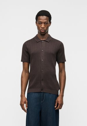 Jeune homme portant une chemise en maille marron foncé à manches courtes boutonnée avec col et un pantalon large bleu foncé, debout devant un fond uni.
