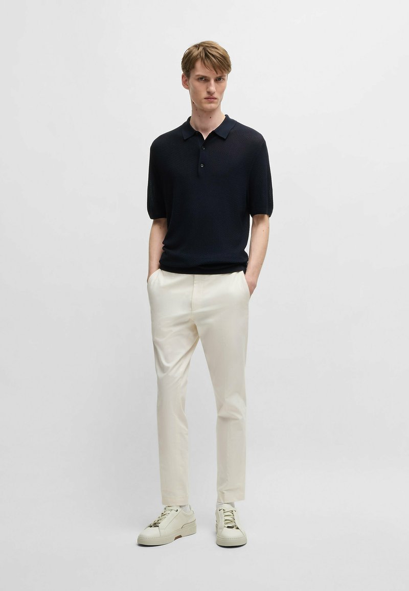 Polo a maniche corte blu navy con motivo testurizzato, abbinato a pantaloni crema e sneakers bianche. Design semplice e moderno.