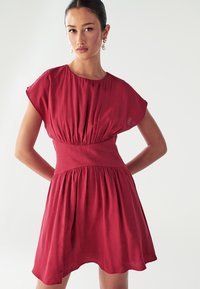 Czerwona sukienka z krótkim rękawem, dopasowana w talii, z lekko rozszerzającą się spódnicą. Wykonana z lekkiej tkaniny o miękkiej strukturze i delikatnych pliskach.