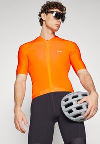 Žiarivo oranžový cyklistický dres s krátkymi rukávmi a zipsom vpredu, doplnený čiernymi ribbovanými cyklistickými šortkami a drží šedú prilbu.