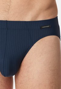 Marineblaue Badebrieft mit vertikalen Streifen und einem schwarzen Etikett. Hergestellt aus dehnbarem Material, mit einem strukturierten Design und glatter Oberfläche.