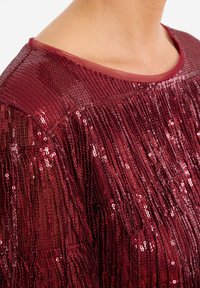 Bordeaux top met een doorschijnende halslijn en franjes van pailletten, met een getextureerd, glanzend oppervlak.