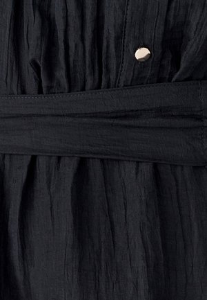 Černá texturovaná tkanina s zvrásněným vzhledem se vyznačuje odpovídajícím páskem a lesklým kovovým knoflíkem u pasu.