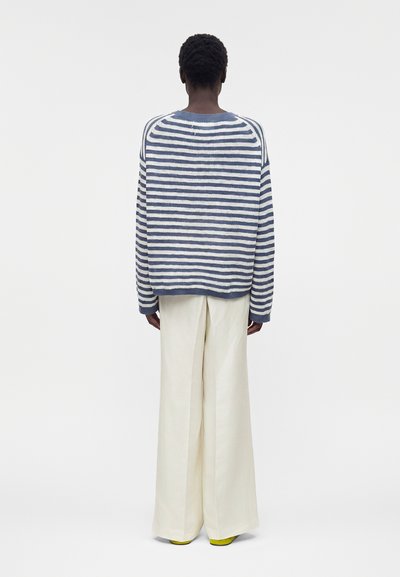 Άτομο με κοντά σγουρά μαλλιά που φοράει μπλε και λευκό ριγέ πουλόβερ, φαρδιά κρεμ παντελόνια και κίτρινα παπούτσια, στέκεται γυρισμένο με την πλάτη.