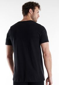 Hombre con cabello rizado que lleva una camiseta negra de manga corta lisa, de espaldas con la cabeza girada hacia un lado sobre un fondo blanco.
