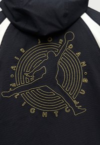 Μαύρο φούτερ με κουκούλα με κίτρινο κεντημένο λογότυπο Jumpman καλαθοσφαίρισης και κυκλικό κείμενο "Original Flight" στην πλάτη.