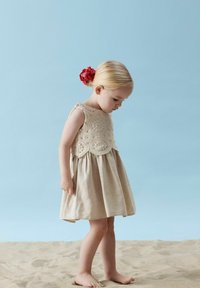 Beiges Leinenkleid mit gehäkeltem Oberteil, gewelltem Saum und ärmellosem Design. Rotes Blumenkopfbedeckung. Steht auf Sand.