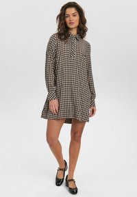 Robe à manches longues à carreaux bruns et noirs avec un col et des boutons, en tissu texturé et avec un ourlet évasé. Des chaussures noires sont portées.