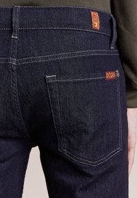 Musta siniset farkut, joissa on takatasku, jossa on broderad design och en kontrasterande röd logotyp på midjan.