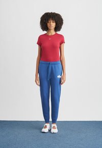 Červená krátkym rukávom košeľa s bielym logom, doplnená modrými teplákami s bielym logom a bielymi športovými topánkami. Jednoduché pozadie.