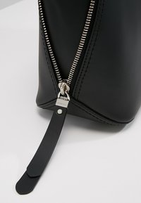 Gros plan d’un coin de sac en cuir noir montrant une fermeture éclair argentée partiellement ouverte avec une longue languette en cuir noir attachée.