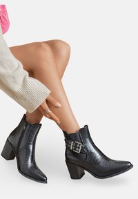 Jambes croisées, portant des bottines en cuir noir texturé avec des talons épais et des sangles bouclées, associées à une manche en tricot beige et un top rose.
