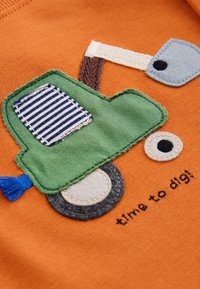 Grüner und blauer Stofftraktor mit aufgenähten Rädern und Auspuff auf orangefarbenem Stoff, begleitet von dem Text „Zeit zum Graben!“