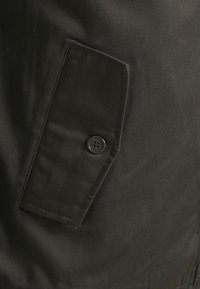 Tissu foncé avec un rabat rectangulaire maintenu par un bouton noir à quatre trous, montrant les coutures détaillées et la texture du matériau.