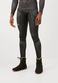 Sorte atletiske leggings med en glat tekstur, prydet med teksturerede mesh-paneler og hvide accentlinjer. Båret med sorte høje sko.
