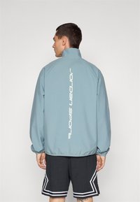 Cazadora cortavientos de color azul claro con un cuello alto, que presenta un logo vertical en blanco de "Jordan Sport" en la parte posterior, llevada con pantalones cortos negros.