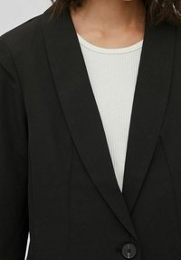 Černý blejzr s klopou a jedním knoflíkem, vyrobený z hladkého materiálu. Nositelný přes světle vroubkované tričko.