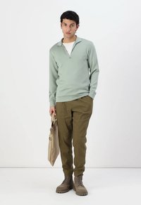 Suéter verde claro con media cremallera, combinado con pantalones verde olivo y botas marrón con cordones. Bolsa beige sostenida en una mano.