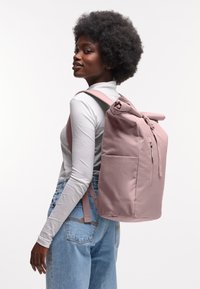 Sac à dos en tissu rose avec fermeture enroulée, zips et poches latérales. Texture lisse et forme structurée, porté de manière décontractée sur l'épaule.