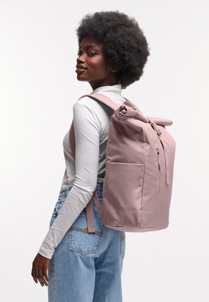 Sac à dos en tissu rose avec fermeture enroulée, zips et poches latérales. Texture lisse et forme structurée, porté de manière décontractée sur l'épaule.
