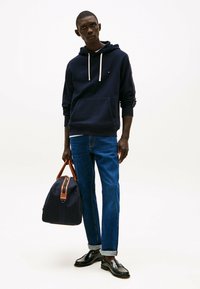 Navy hoodie met trekkoorden en een voorkeurszak, gecombineerd met blauwe spijkerbroeken en zwarte loafers. Houdt een donkere tas met bruine accenten vast.