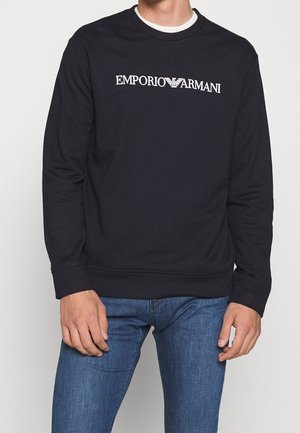 Homme portant un sweat-shirt noir Emporio Armani avec un logo blanc et un jean bleu sur un fond uni.