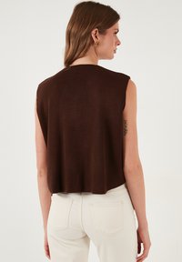 Brązowy bezrękawnik wykonany z prążkowanego materiału, o luźnym kroju i zaokrąglonym dolnym brzegu, zestawiony z jasnoskalowymi, biodrowymi dżinsami.