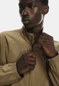 Helle braune Zip-Jacke mit hohem Kragen. Verfügt über elastische Bündchen und Ton-in-Ton-Logo-Details. Glatte Textur auf leichtem Stoff.