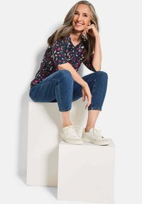 Marinebluse mit floralem Muster, kombiniert mit blauen Jeans und weißen Sneakern; Modell sitzt auf einem weißen Podest und präsentiert einen lässigen Stil.