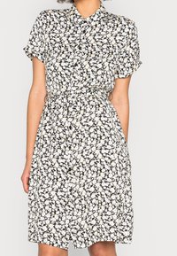 Svart klänning med vitt och gult blommönster, korta ärmar och knäppning. Har en insvängd midja för en definierad form.