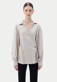 Ljusgrå omlottsblus med spetsig krage, lös passform och sidoknappdetalj, kombinerad med ribbade svarta byxor. Slät tygstruktur.