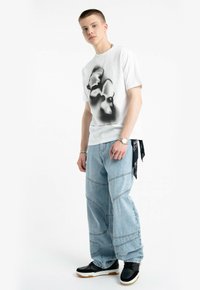 Biała koszulka z czarnym nadrukiem graficznym, zestawiona z jasnoniebieskimi dżinsami o szerokich nogawkach oraz czarnymi trampkami z białą podeszwą.