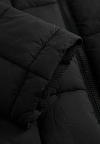 Detal puchowej, czarnej kurtki z pikowaną teksturą, widocznym przeszyciem i podszytym zamkiem błyskawicznym, prezentujący gładkie, matowe wykończenie.