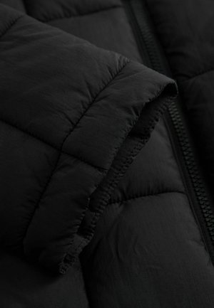 Melnas pūtītas virsjaka detaļas ar šūtu faktūru, redzamām šuvēm un oderētu rāvējslēdzēju, kas demonstrē gludu, matētu virsmu.