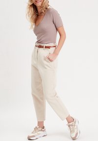 Haut côtelé marron clair avec col en V, pantalon beige taille haute, et baskets beige avec des accents blancs. Une ceinture marron ajoute du contraste.