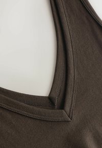 Prim-plan al unui maiou maro din țesătură, care arată decolteul pătrat cu dublă cusătură și bretelele pe umăr, pe un fundal deschis la culoare.