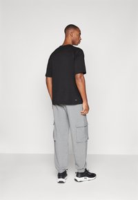 Mężczyzna ubrany w czarną koszulkę z krótkim rękawem, szare spodnie dresowe typu cargo i czarne tenisówki, stojący tyłem na jednolitym białym tle.