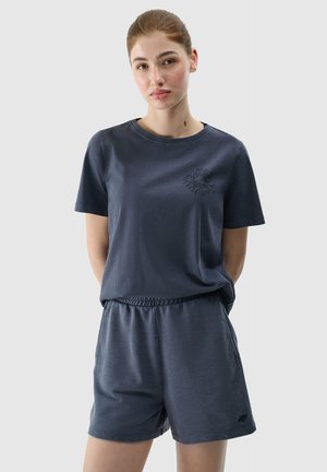 Młoda kobieta z włosami związanymi do tyłu, ubrana w ciemnoniebieski t-shirt z krótkim rękawem i pasujące szorty, na jednolitym jasnym tle.