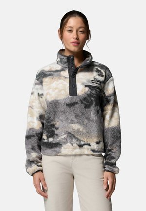 Młoda kobieta ubrana w szarobeżowy polar z wzorem, z zatrzaskami i kieszenią na piersiach zapinaną na zamek, stojąca na tle jednolitego tła.