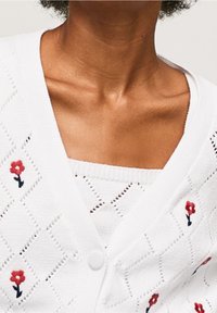 Bílý pletený cardigan se vzorem diamantů, zapínáním na knoflíky a červenou květinovou výšivkou. Měkká textura s detaily otevřeného úpletu.