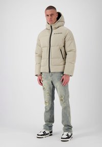 Beige gewatteerde pufferjas met een rits, voorzakken en een gewatteerd ontwerp. G gedragen met lichte distressed jeans en gekleurde sneakers.