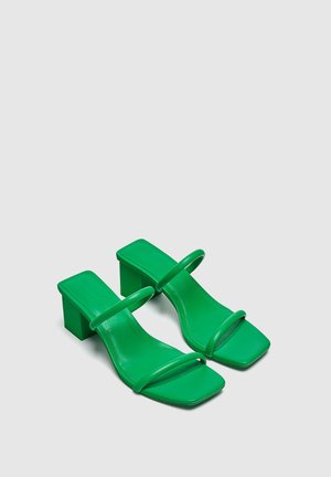 Par de sandalias verdes de tacón con punta cuadrada, tacón ancho y dos finas tiras sobre el pie, sobre un fondo claro y liso.