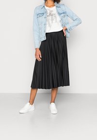 Chaqueta de mezclilla azul claro sobre una camiseta gráfica blanca, combinada con una falda midi plisada negra y zapatillas blancas. Texturas suaves y diseño clásico.
