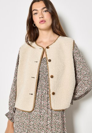 Gilet in pile beige con scollo rotondo, rifinito da bordi marroni e bottoni in legno. Indossato sopra un vestito con stampa floreale.