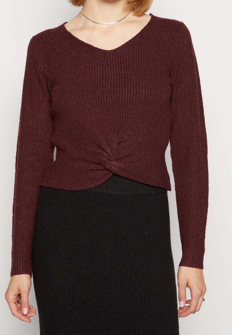 Bordeaux ribben gebreide trui met een V-hals en geknoopte details bij de taille, gecombineerd met een zwarte ribben bodycon rok.