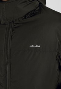Černá zateplená bunda s vysokým límcem, předním zipem a bílým logem "night addict" na levé části hrudi. Hladký povrch.