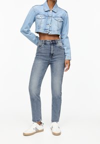 Personne portant une veste courte en denim bleu clair, un jean bleu délavé taille haute et des baskets blanches avec des semelles en gomme, debout devant un fond blanc.
