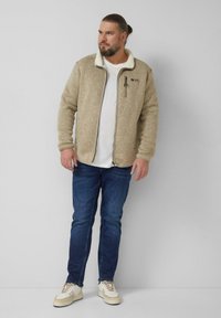 Veste en polaire beige avec col montant, dotée d'une poche zippée et d'une surface texturée. Portée sur un t-shirt blanc et un jean bleu.