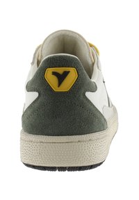 Zapatilla blanca con acentos de gamuza verde, con una entresuela texturizada color crema y un detalle del logo amarillo en el talón. Diseño limpio y minimalista.