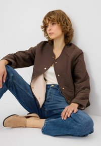 Giacca trapuntata marrone con un motivo geometrico, interno crema e chiusure a scatto dorate, abbinata a jeans blu a vita alta e mocassini beige.
